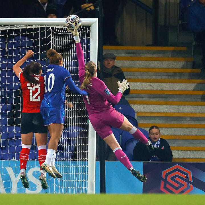 Soccer goalkeeper in pink diving to save a ball, with teammates and audience in the background.